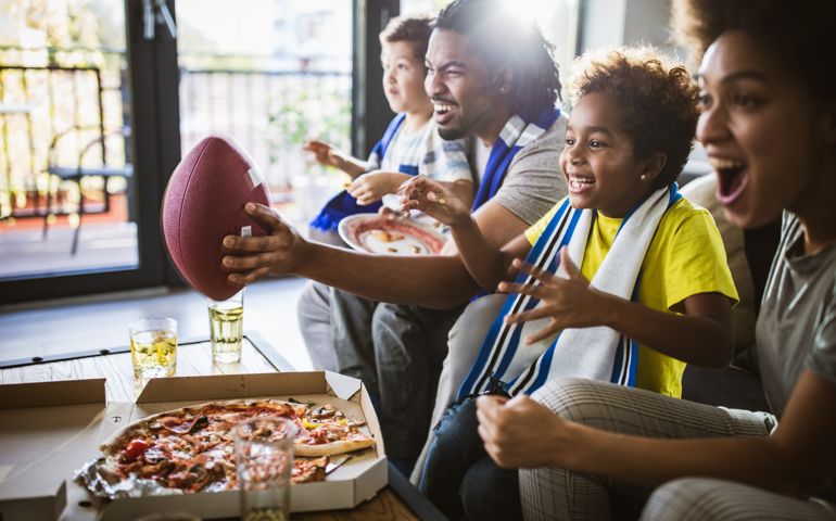 Young joyful black family having fun while cheering for their favorite American football team at home.