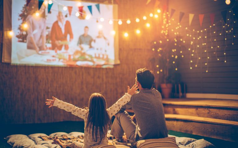 Father and daughter sitting at backyard and looking movie at home improved theatre. Backyard is decorated with string lighs.