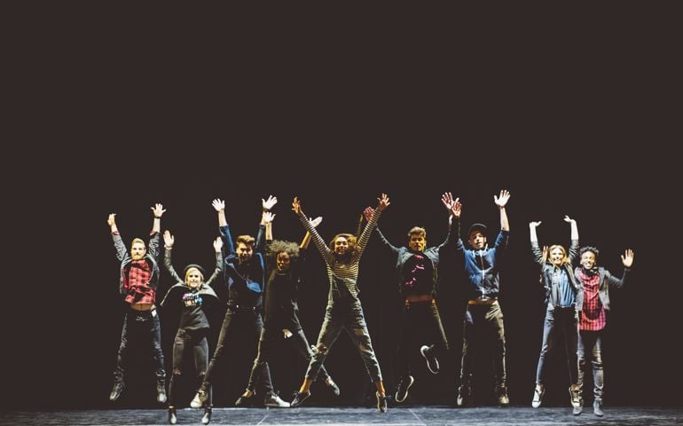 Group of young performers on the stage standing in the line, jumping with raised hands. Dark tones.