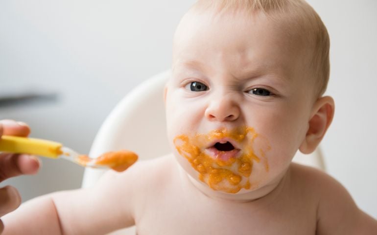 Mother feeding messy baby son with spoon in high chair