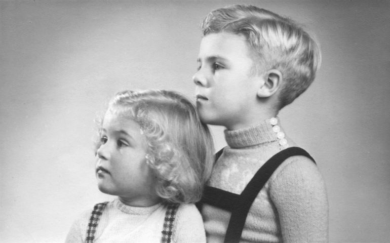 Early 1950s duo portrait of a young boy and girl with blond hair and curls looking sideways.