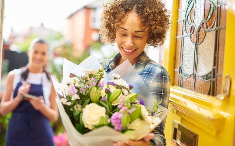 A young woman stands at the front door of her home and reads the card from a big bouquet of flowers . Behind her is the delivery girl who has just dropped them off. The shallow focus is on the bouquet blurring off to the people.