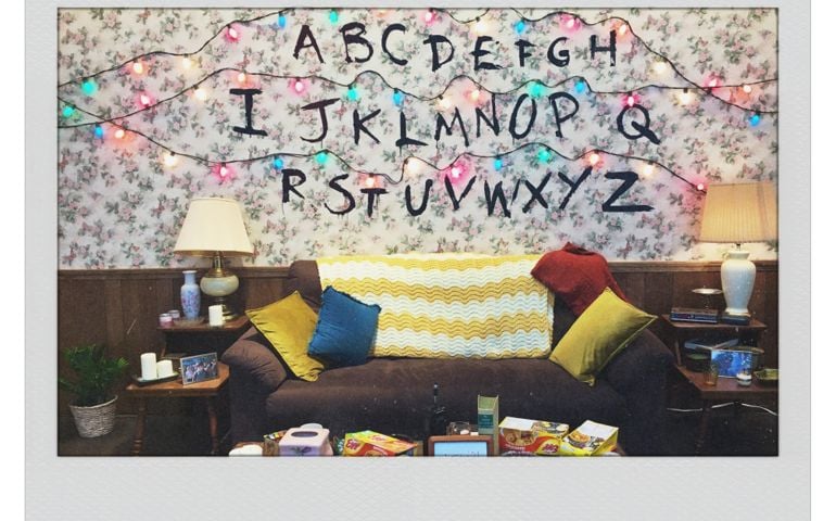 A living room setup features floral wallpaper with the alphabet painted on it in black letters and colorful Christmas lights strung above each row. The lights are lit in various colors, referencing a scene from Stranger Things. A brown couch with throw pillows and blankets sits below, surrounded by lamps, books, and scattered snacks.