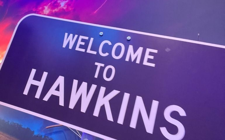 A tilted road sign reads “Welcome to Hawkins” in bold white letters, set against a dramatic purple and red stormy sky with lightning. The scene references the fictional town in Stranger Things.