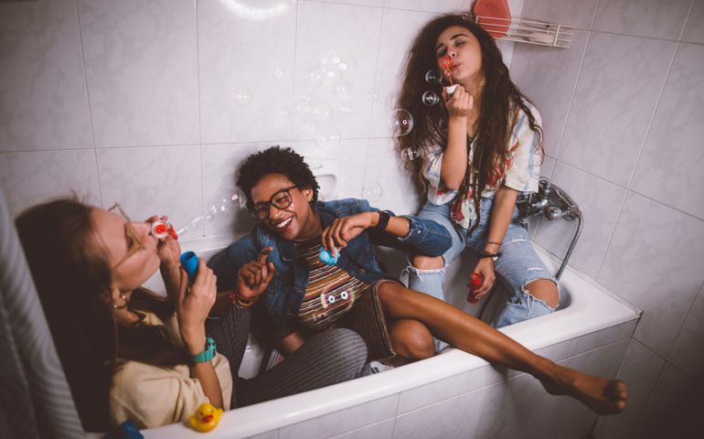 Young multi-ethnic hipster girls having slumber party at home and blowing bubbles in bathtub