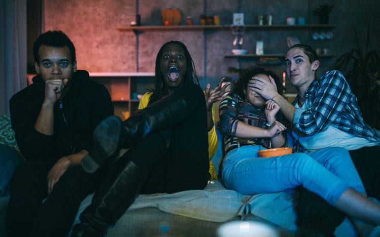 Young friends watching horror movie on TV at home, eating popcorn and chips at night