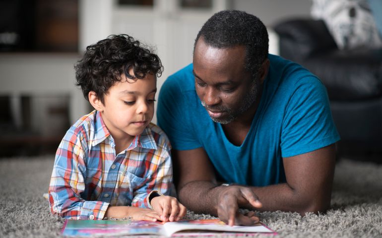 A young father of African descent is lying on the carpet in their living room and reading to his son.
