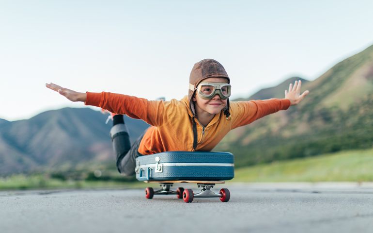 A young boy wearing flying goggles and flight cap has packed his suitcase and has placed it on a skateboard. He is ready to fly away with arms outstretched to the destination of his dreams. Image taken in Utah, USA.