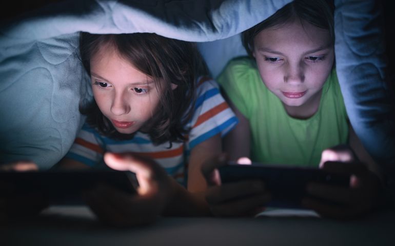 Boy and girl playing games on mobile phones while lying on bed in bedroom under the blanket. They are spending some nice time together that makes them happy.