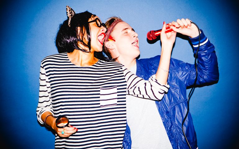 Portrait of a young man and woman in fancy dress singing into a microphone standing in front of a blue backdrop.