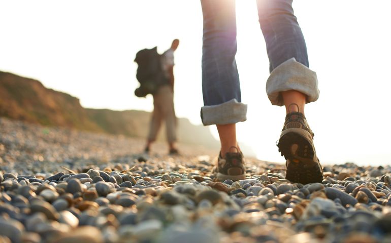 Shot of woman's shoes as a couple walk along a pebble beach towards afternoon sun. Man out of focus in background wearing rucksack.