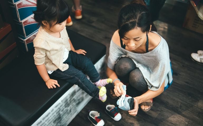Pretty young mom helping toddler girl fitting with a new pair of shoes in a shoes store.