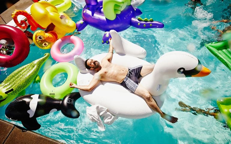 Smiling man relaxing on large inflatable swan in outdoor pool filled with multiple pool toys and friends swimming in background