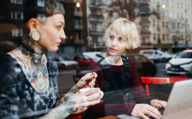 A tattooed and a blonde young woman are talking to each other inside a bright café. One if them is typing something on a notebook while the other one is holding a cup of coffee in her hands.