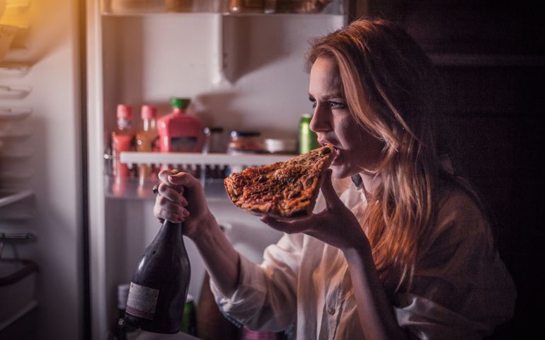 Young woman having vice, eating pizza and drinking wine late night in front of the refrigerator