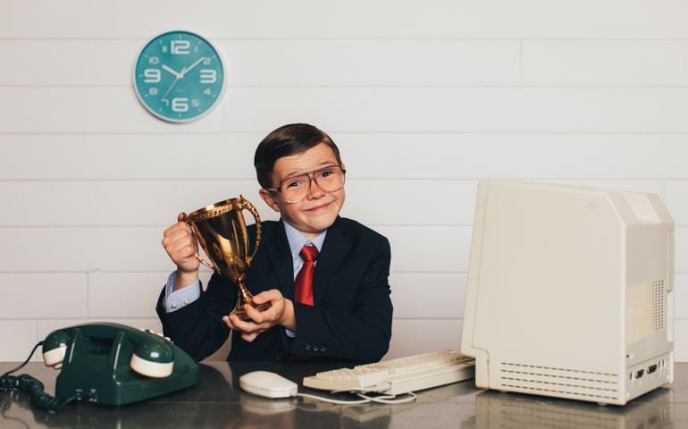 A young business boy dressed in business suit and tie sits at his office desk holding his winning trophy. He is super hard worker and has recently won every competition in company performance.