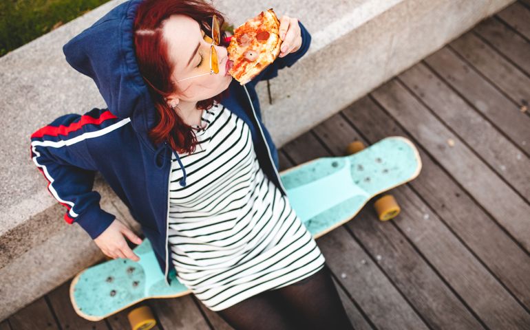 High angle view of young skater girl having a slice of pizza outdoor, sitting on skateboard