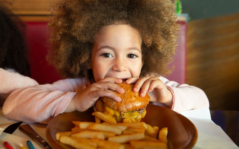 Ethnic kid girl eating burger and chips hungry at indoor restaurant