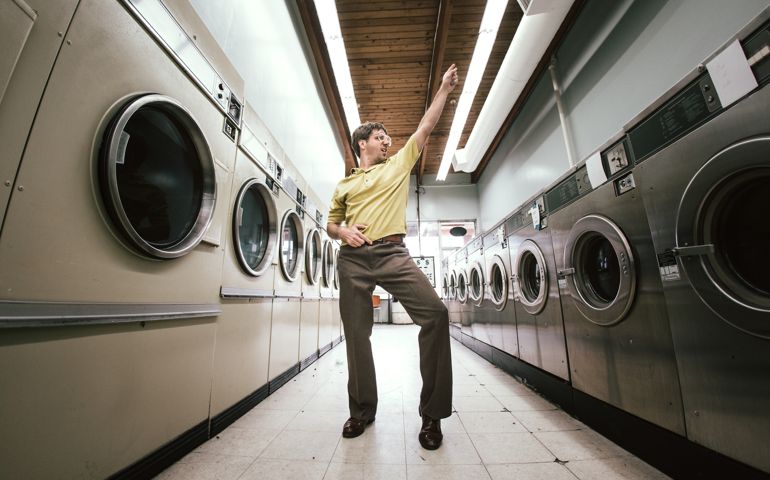 A man in 1980's style does his laundry at an old school laundromat, dancing disco style in the walkway between the washers and dryers while waiting for his clothing to finish drying. Horizontal fisheye image with copy space.