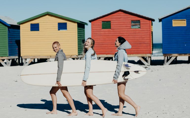 Three young women laughing and smiling while walking with a long board on the beach in front of some colored houses