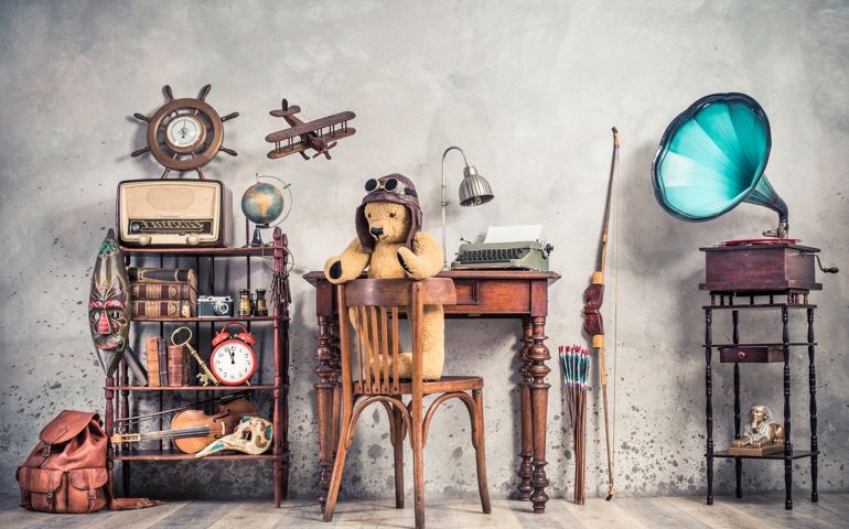 Teddy Bear toy on chair, typewriter, vintage gramophone, old books, radio, globe, binoculars, carnival mask, camera, fiddle on shelf, steering wheel, plane, travel backpack, bow. Retro style photo