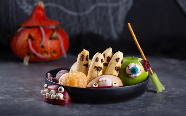 Banana ghosts with chocolate faces, clementine pumpkins and apple monsters on black plate. Fruits in Halloween decoration on dark background.