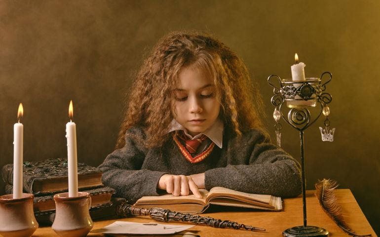 Portrait of a cute little witch girl with magnificent long brown hair dressed in a navy blue jumper, white shirt and red tie. She is reading a book with spells while sitting at the table against a black smoky studio background. There are magic wand, candles in candlesticks on it. The concept of magic, wizardry, miracles. Close-up.