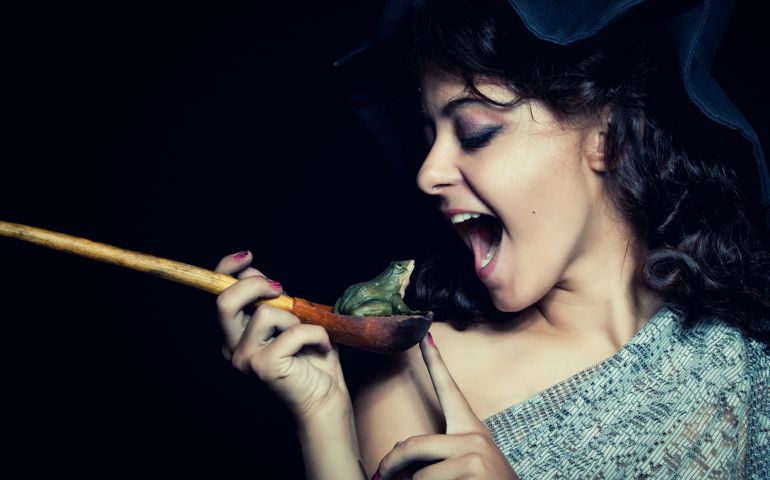 Pretty witch in hat eating frog over dark background