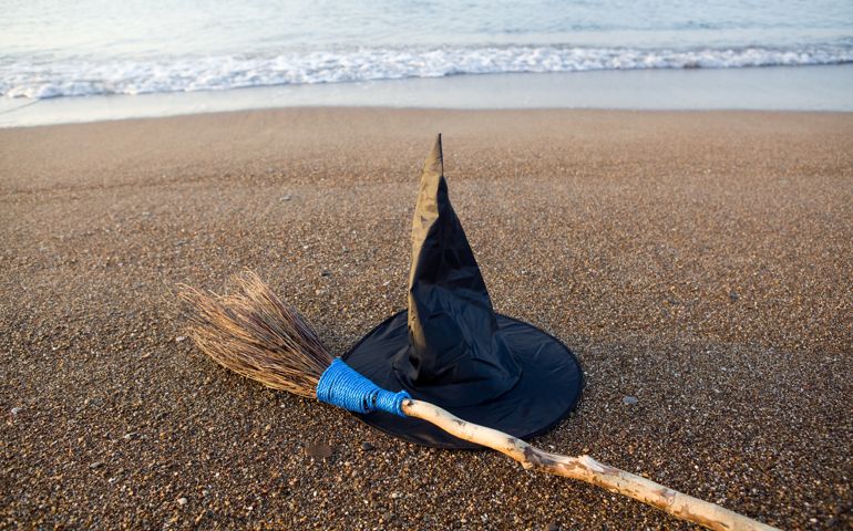Photo of witch's hat and broom on sands in the beach by mediterranean sea.Sea waves are seen on top of frame.the broom is located on left down diagonal position.No people seen in the frame.The photo was taken by a full frame DSLR camera in daylight.