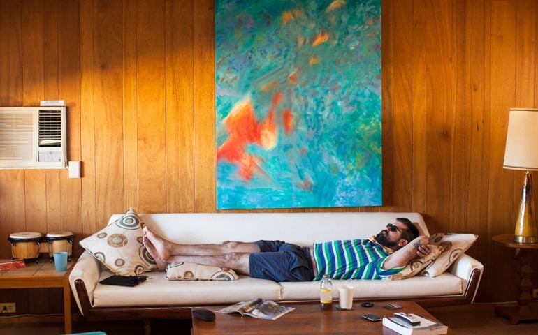 Bearded man with tattoos wearing sunglasses laying on a white sofa under an abstract painting taking a nap