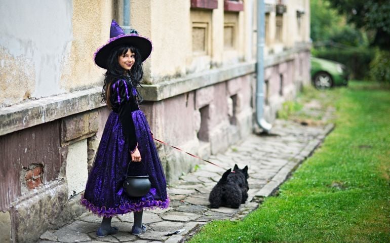 Little girl during halloween dressed up as a witch walking with a dog by old weathered building. The girl is aged 9 and is carrying a little cauldron for candy. The girl is smiling over shoulder at the camera.