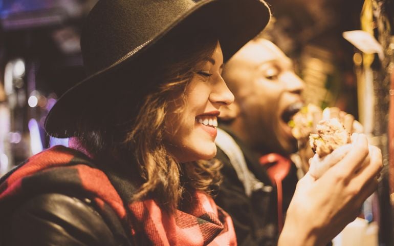 Beautiful young woman wearing black hat eating burger in the pub. Close up of face. Dark tone.