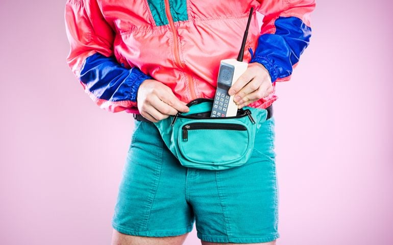 A man wearing fluorescent colored clothing puts a 1980's - 1990's cellular brick phone into his fanny pack, representing state of the art style and technology for that time. Detail shot; horizontal with pink background.