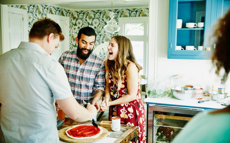 Laughing couple preparing pizza together with friends in kitchen