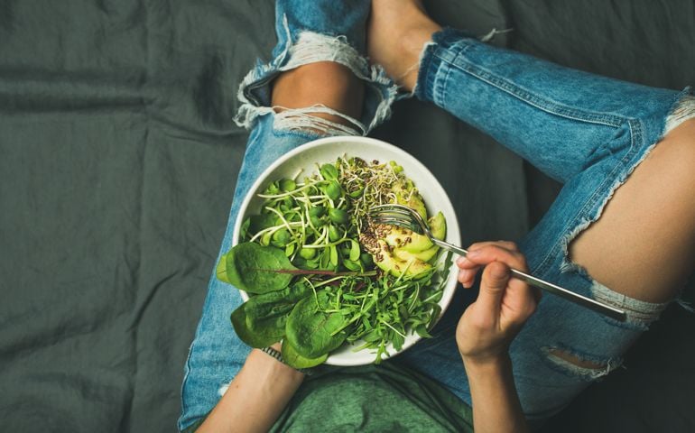 Green vegan breakfast meal in bowl with spinach, arugula, avocado, seeds and sprouts. Girl in jeans holding fork with knees and hands visible, top view, copy space. Clean eating, vegan food concept