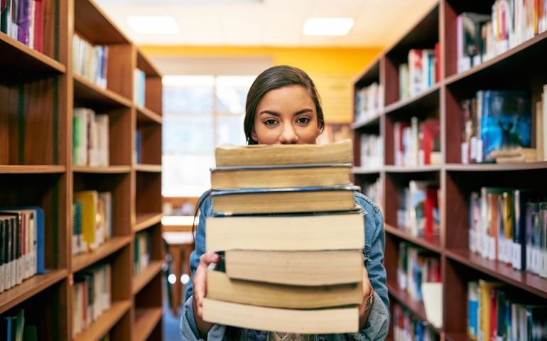 Portrait of a university student holding a pile of books in the library at campus