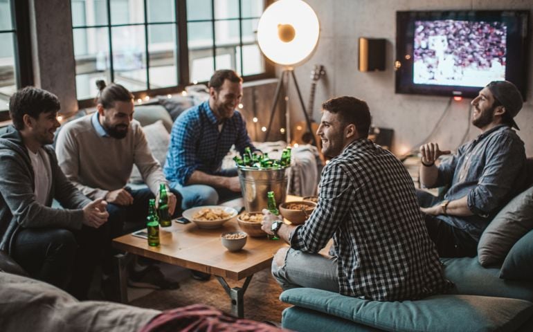 Group of young men gather at home party. They are watching basketball game on tv, drinking beer, making jokes and cheer for their team.