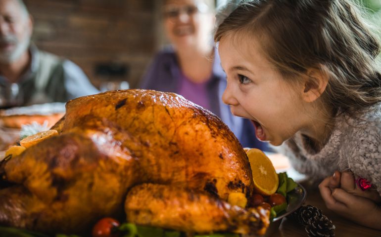 Cute little girl having fun while about to bite a stuffed turkey during Thanksgiving dinner in dining room.