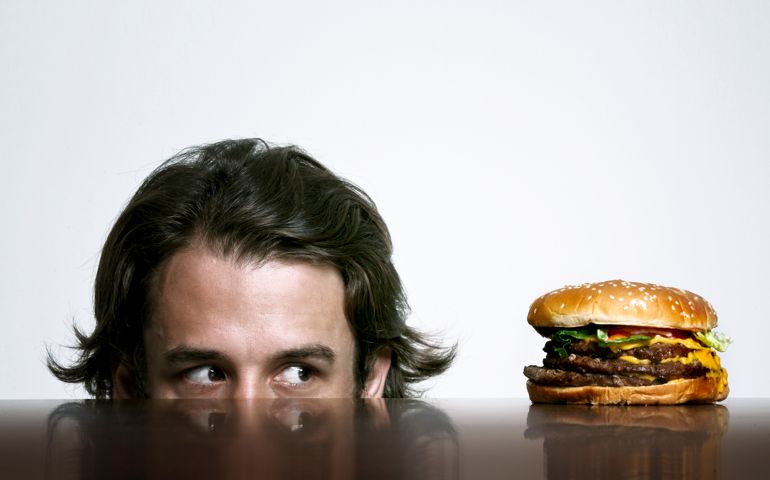 a guy looking at a huge hamburger like he is coming from below a table