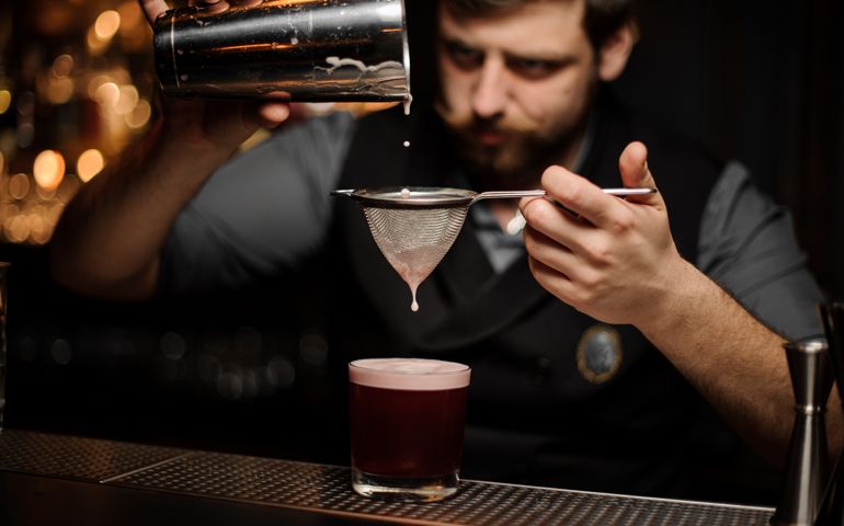 Bartender with a beard pouring a smooth pink cocktail through the sieve to the glass from the shaker on the bar counter on the blurred background.