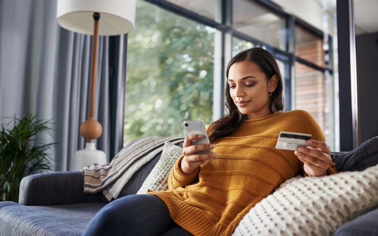 Shot of a beautiful young woman using her cellphone and credit card while relaxing on a couch at home