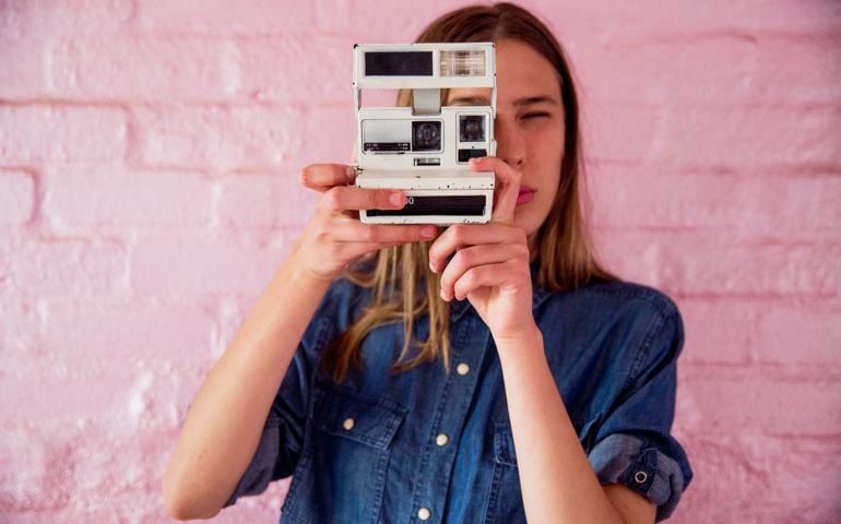 Beautiful young woman on pink backdrop taking a photograph with retro camera