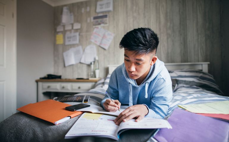 Teenage boy lying on his bed while concentrating on homework for his exams.