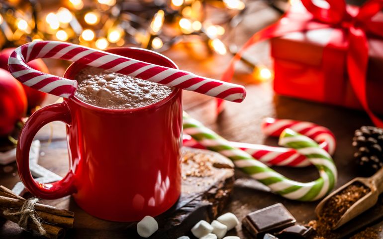 Red mug filled with homemade hot chocolate shot on rustic Christmas table. A red and white candy cane is on the hot chocolate mug and two others candy canes are behind the mug. String Christmas lights and a red gift are out of focus on background. Predominant color is red. DSRL studio photo taken with Canon EOS 5D Mk II and Canon EF 100mm f/2.8L Macro IS USM.
