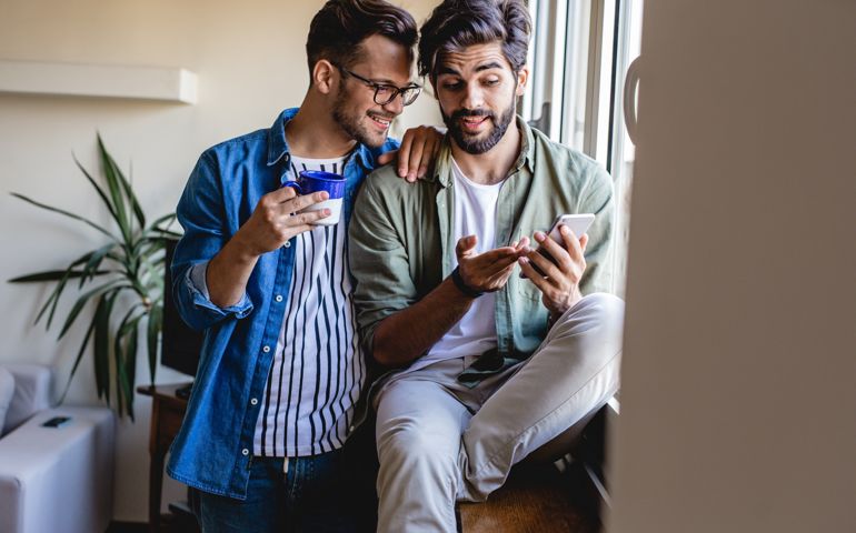 Gay men couple at home sit by the window and look at screen of the smartphone