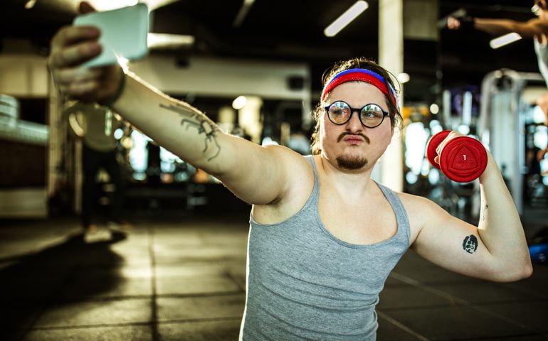Nerdy athlete showing off while exercising with dumbbell in a gym and taking a selfie with smart phone.