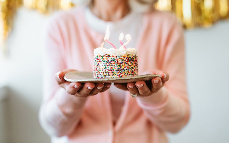 Midsection of retired woman holding cake with burning birthday candles. Senior female is standing with dessert in plate. She is celebrating at home.
