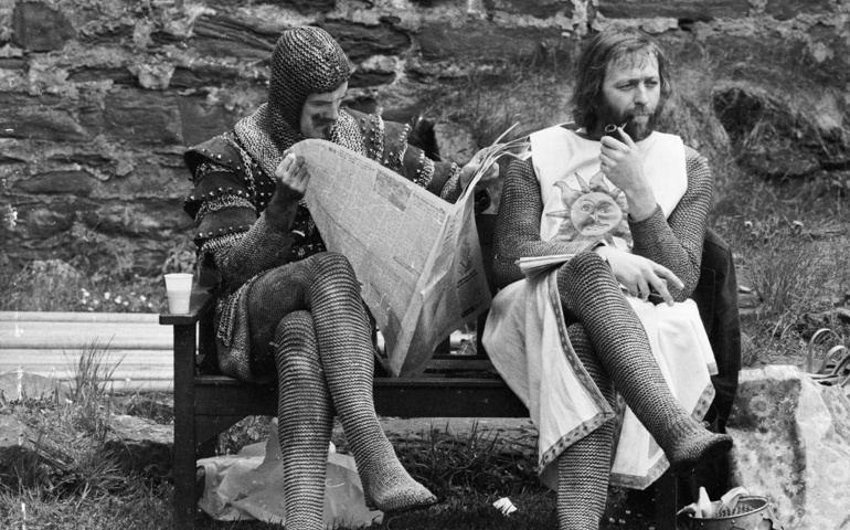 21st May 1974: A chainmail-clad John Cleese reads a newspaper while Graham Chapman smokes a quiet pipe on the set of 'Monty Python and the Holy Grail'.