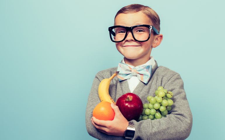 A young nerd boy with glasses is excited to eat his fruit. He is holding a bunch of fruit and has an excited expression on his face.