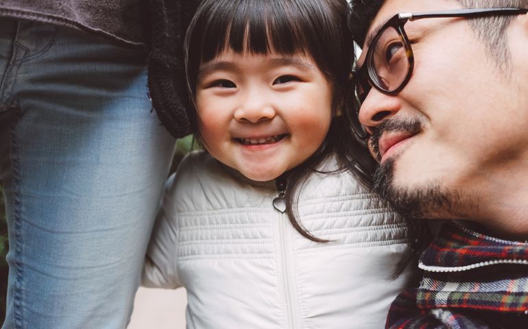 Lovely little girl looking at the camera joyfully with one arm hugging mom's leg &amp; another arm hugging dad's shoulder.